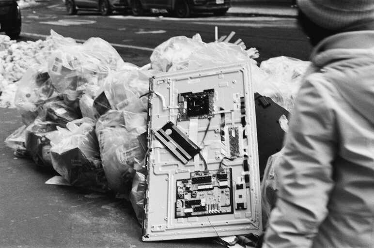 a white metal sheet with attached boards and wiring rests,
          discarded, against white trash bags and snow piled up on a new york city sidewalk
